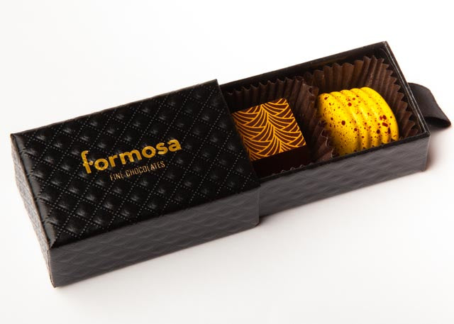 A box of two chocolates, one yellow with brown speckling and one brown with yellow pattern printed on top, presented in a black, textured box with the brand name 'formosa' on the lid.