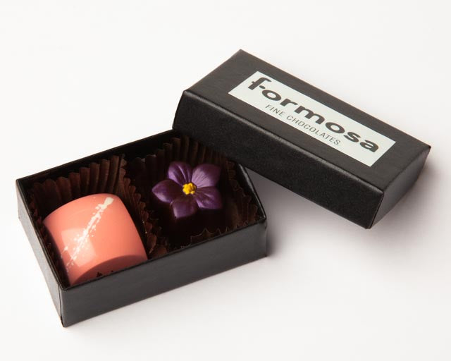 A black box with the Formosa Chocolates logo open to reveal two colorful chocolate bonbons inside.