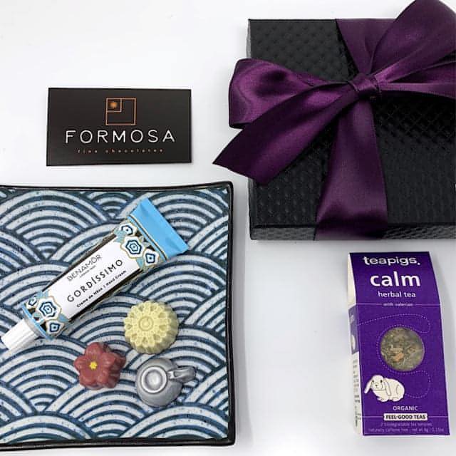 A gift set arranged with a variety of items including chocolates, a box of tea, hand cream, and other small personal care products, all displayed on a decorative plate with a black and purple bow.