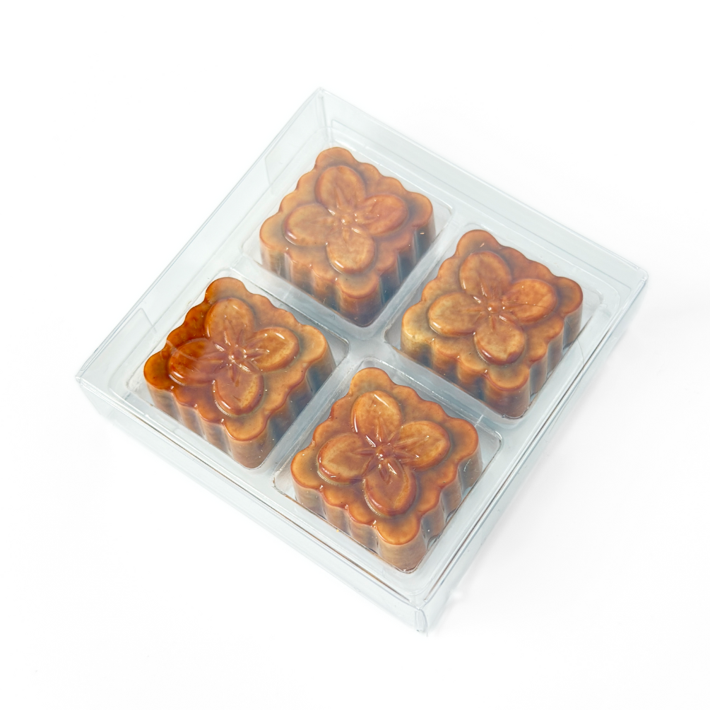 Four square-shaped black sesame praline bonbons with a white chocolate shell and a dark chocolate cap, arranged in a clear packaging on a white background. The product is shaped to look like a traditional moon cake. 