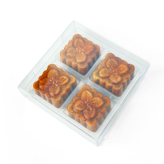 Four square-shaped black sesame praline bonbons with a white chocolate shell and a dark chocolate cap, arranged in a clear packaging on a white background. The product is shaped to look like a traditional moon cake. 